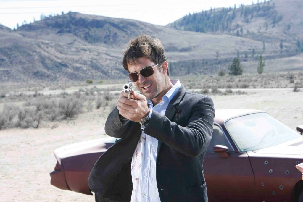 John Sheppard (Joe Flanigan) in the desert with shades on and a bullet-riddled car behind him points his pistol at the camera.