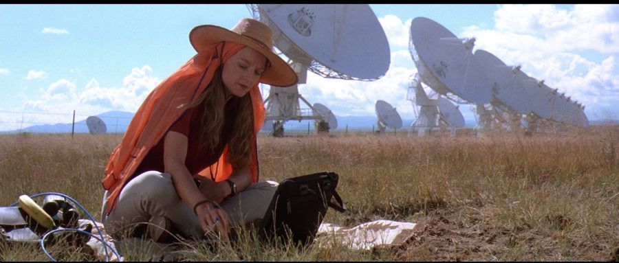 Dr. Ellie Arroway (Jodie Foster) sits on the grass with a row of enormous radio telescopes visible behind her.