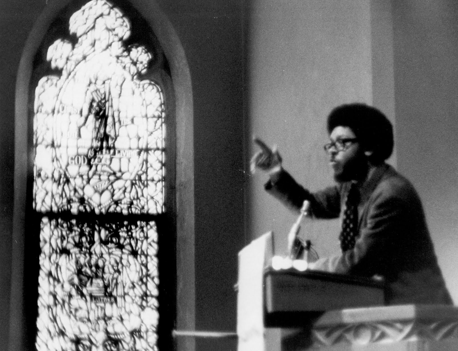James H. Cone speaks at a church pulpit. He is wearing a suit and has glasses and an afro.