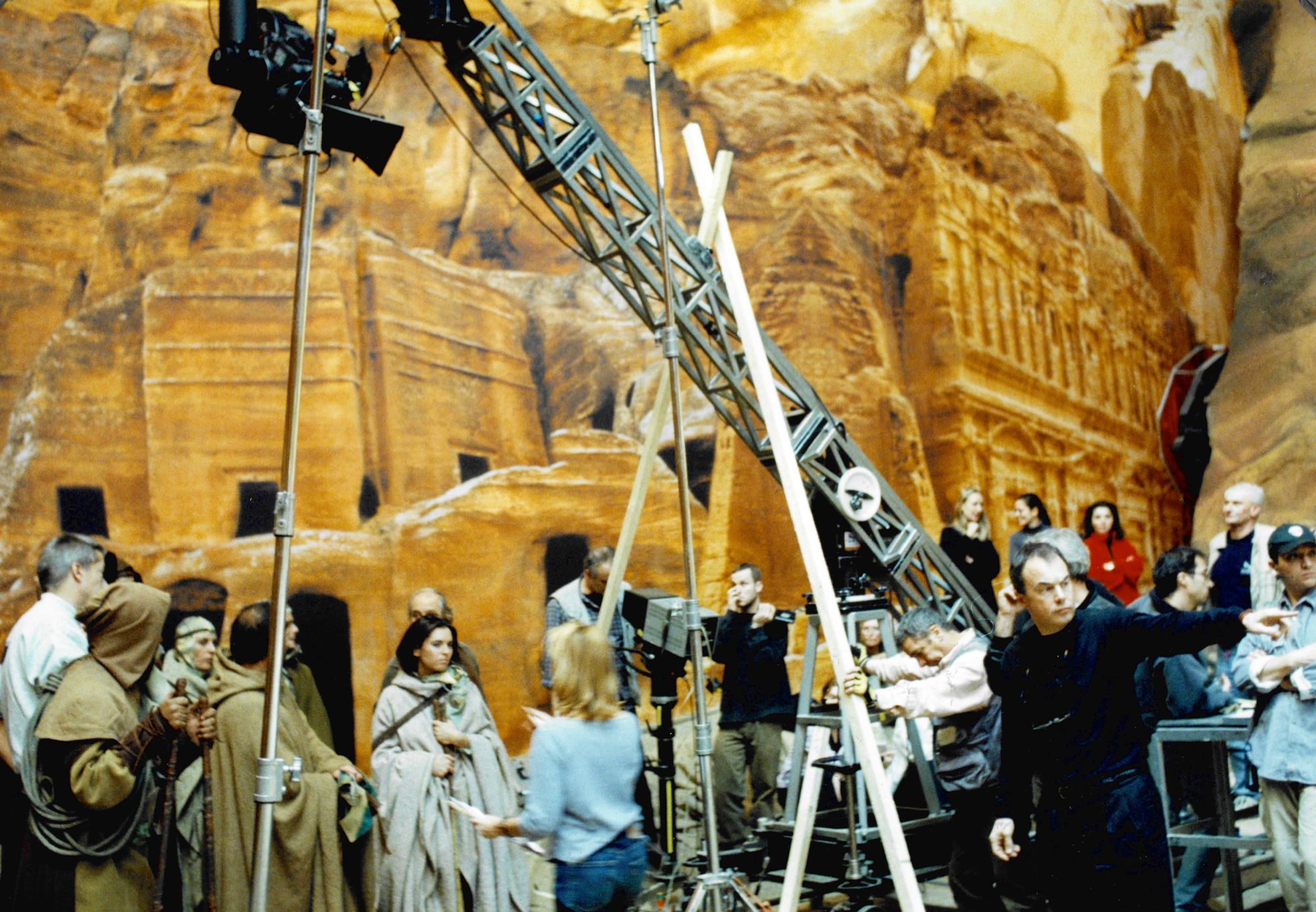 Director John Harrison, wearing b lack, points off camera. Behind him are extras in desert garb and a backdrop showing dwellings cut into a desert rockface.