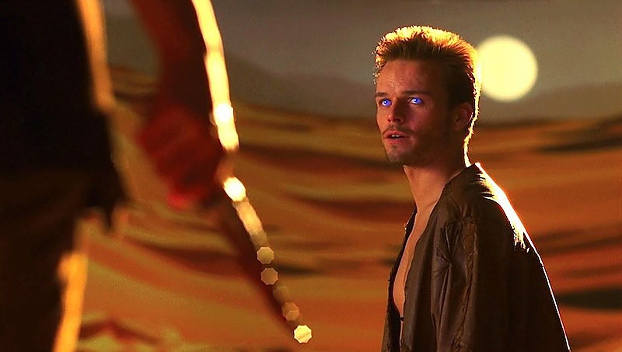 Alec Newman as Paul Atreides looks towards someone who is only partially visible in the foreground. Behind him is the desert and the setting sun.