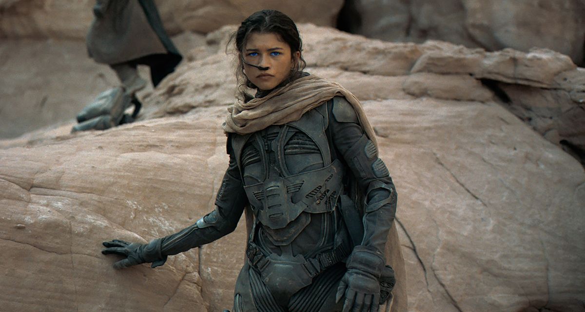 Chani (Zendaya) wearing a stilsuit, rests her hand against a rock.