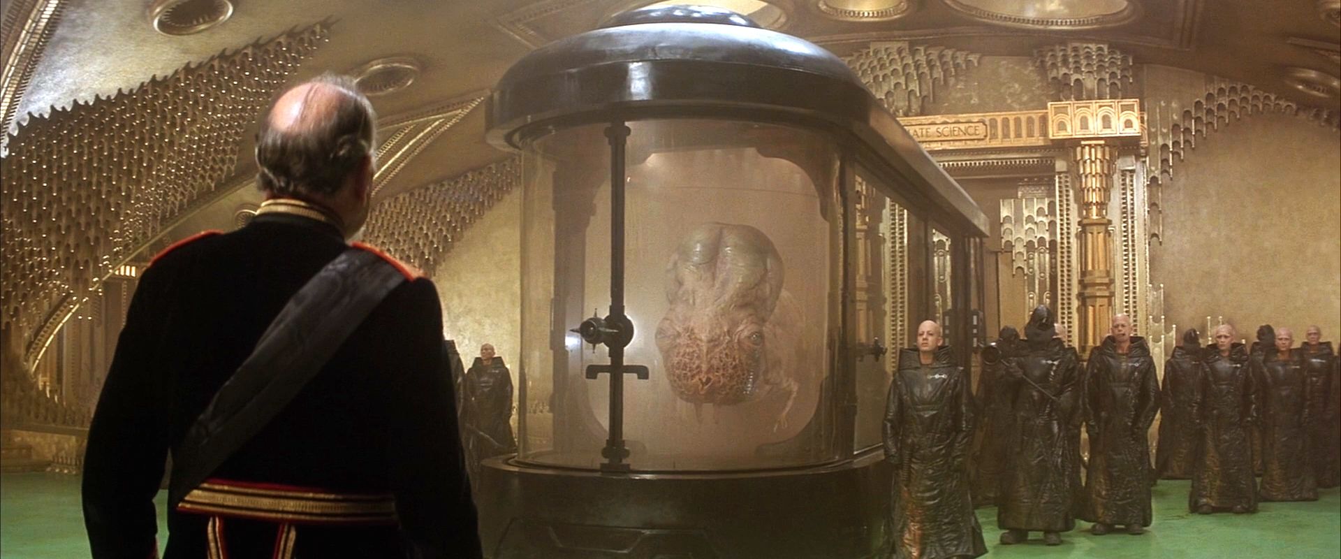 The bulbous, slug-like Guild Navigator floats in a tank flanked by black-robed Guildsmen. The emperor, wearing a black tunic with red and gold accent stands in front of it.