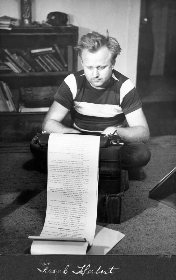 Frank Herbert sits on the floor behind a typewriter. He is wearing a striped T-shirt.