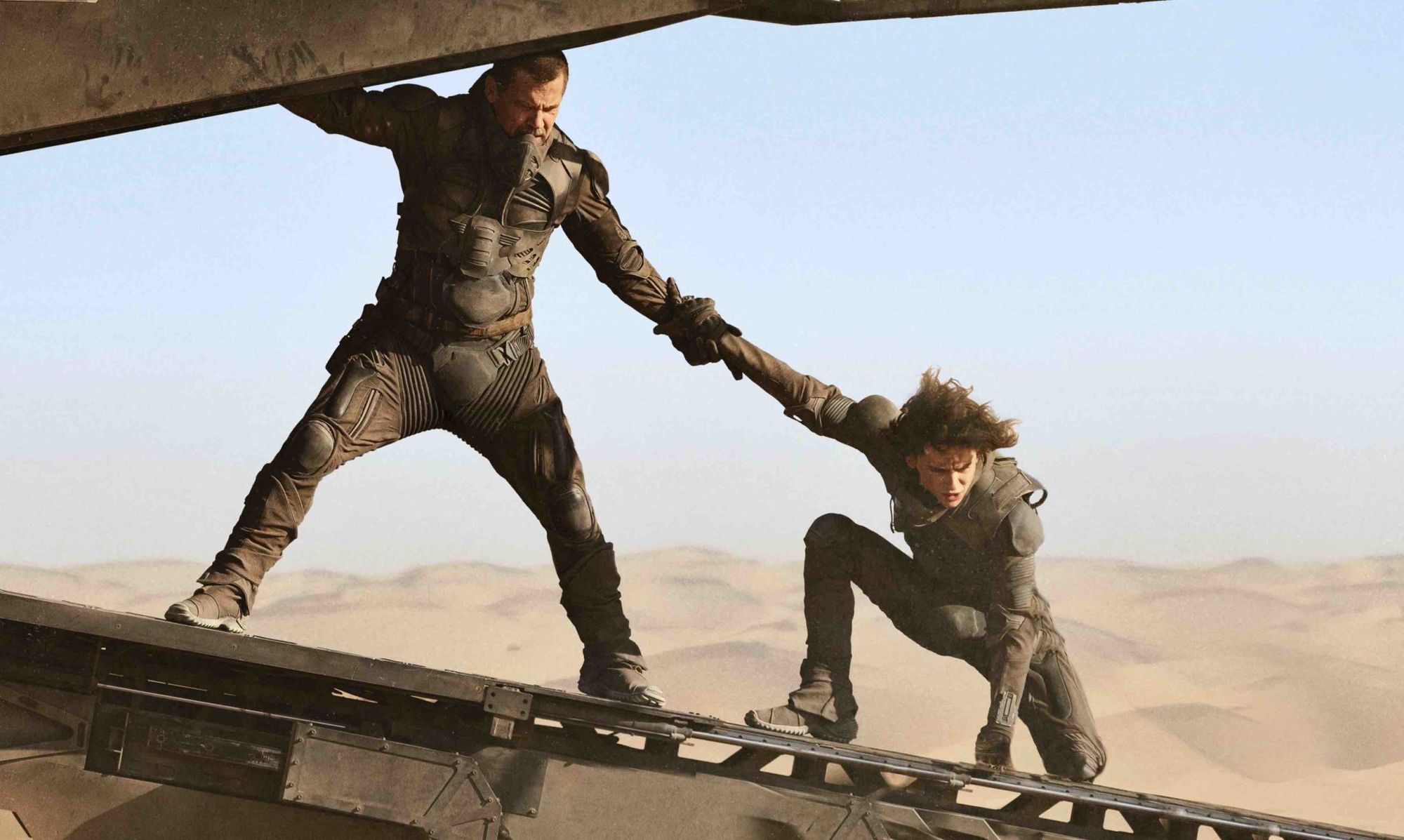 Gurney Halleck (Josh Brolin) and Paul Atreides (Timothée Chalamet) stand on the boarding ramp of an ornithopter in flight, the desert behind them.