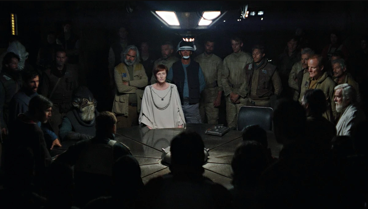 Mon Motha (Genevieve O'Reilly) stands at a round table in a darkned room, surrounded by men in olive military fatigues.