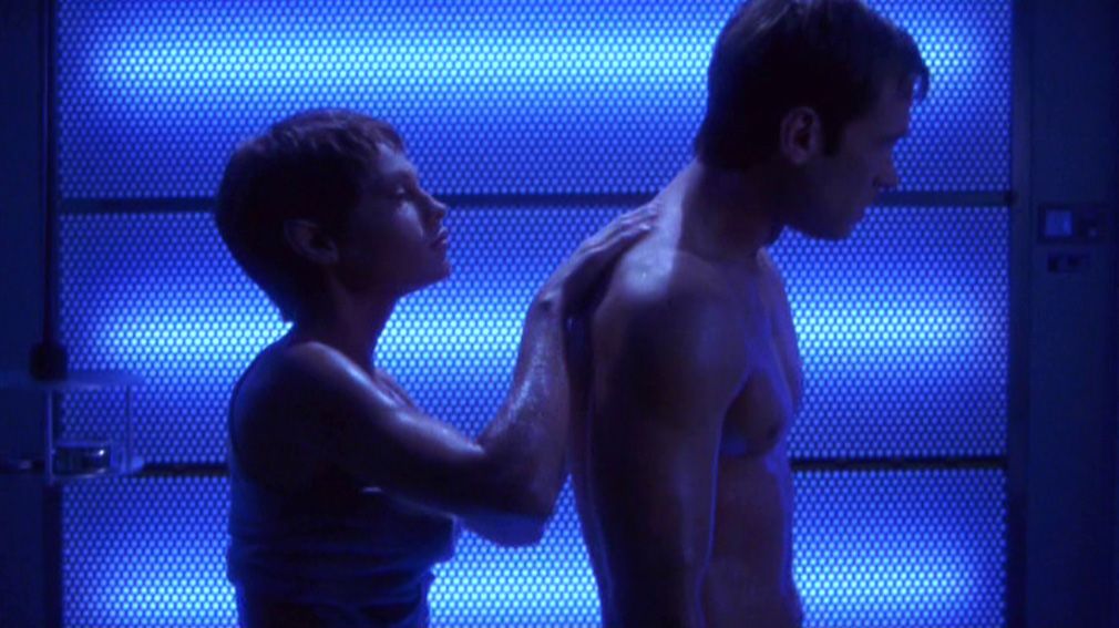 T'Pol (Jolene Blalock) rubs gel into Tucker (Connor Trinneer)’s naked back, backlit by blue lights.