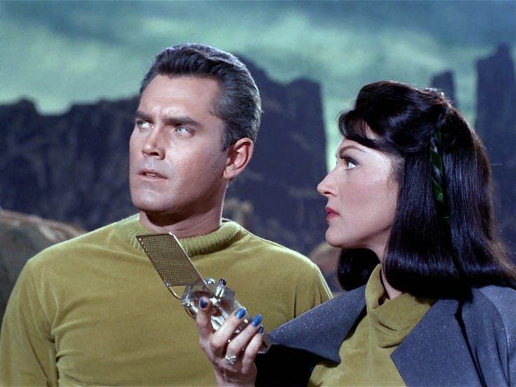 Captain Pike (Jeffrey Hunter) and Number One (Majel Barrett) on an alien world. Number One holds up a communicator.