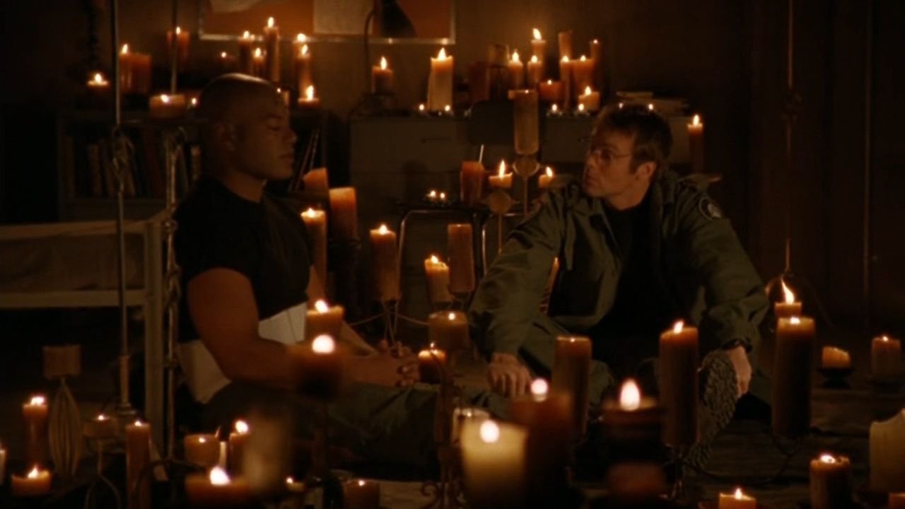 Teal'c (Christopher Judge) and Daniel Jackson (Michael Shanks) sit on the floor surrounded by candles. Teal’c has his back against the bed frame and his eyes closed.