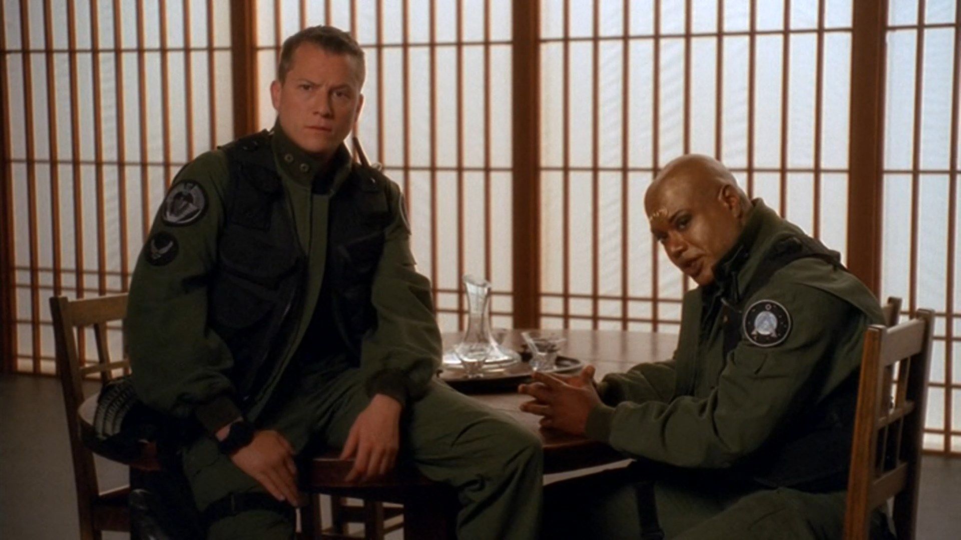 Jonas Quinn (Corin Nemec) sits on a table and Teal'c (Christopher Judge) sits on a chair next to him.