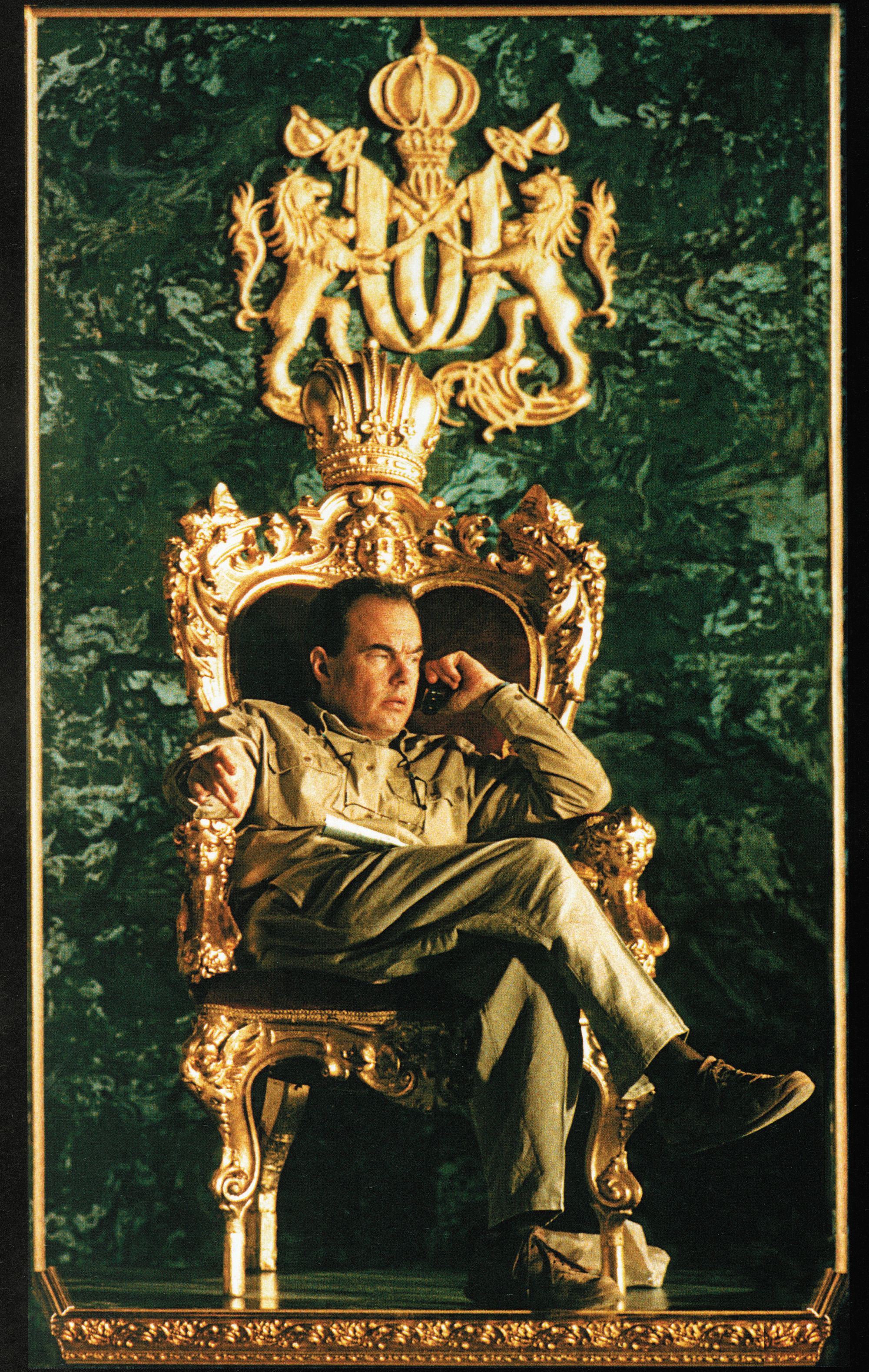 Director John Harrison is wearing tan colored shirt and slacks. He sits on a gold chair with a high back and listens to someone on a mobile phone. The wall behind him is green marbel.