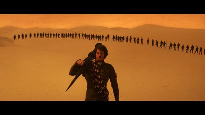 Paul Atreides (Kyle MacLachlan) in a stillsuit walks across the desert with a thumper and some rope over one shoulder.