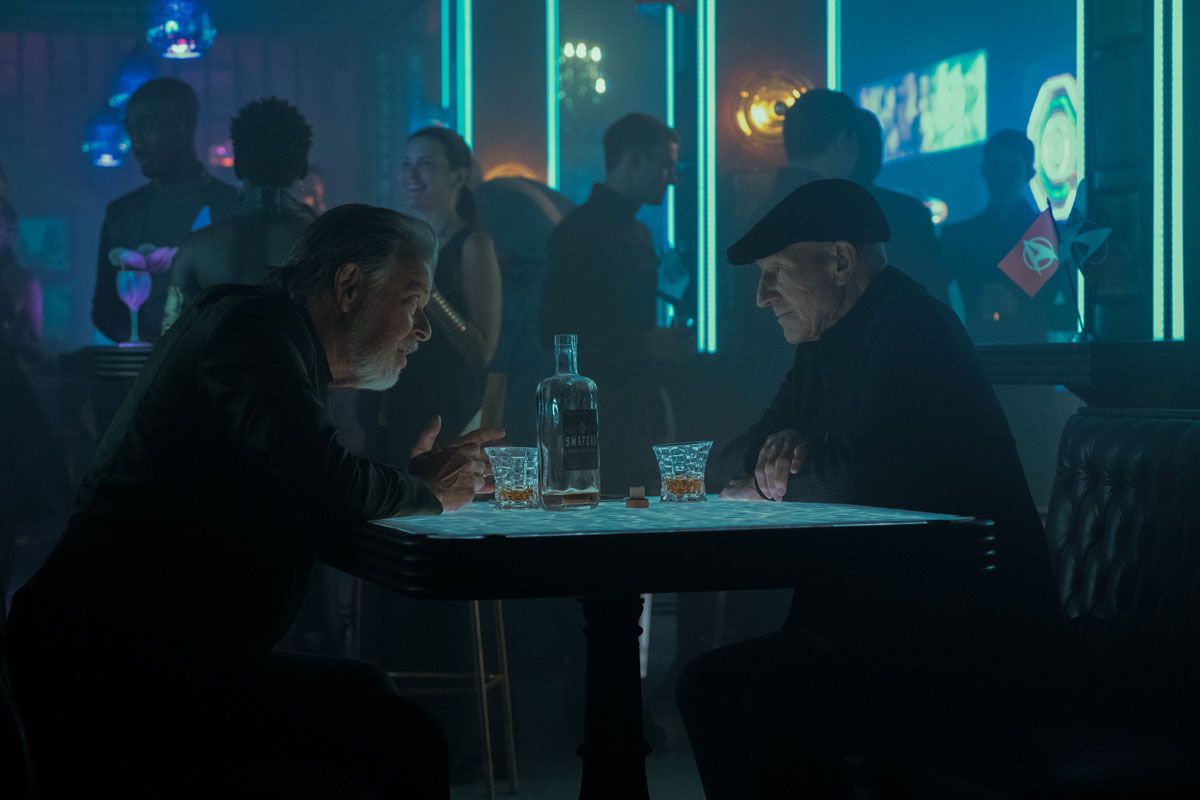 Will Riker (Jonathan Frakes) and Jean-Luc Picard (Patrick Stewart) sit either side of a table in a dimly lit bar. There's a bottle of whisky between them.