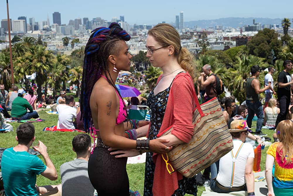 Nomi Marks (Jamie Clayton) and her girlfriend Amanita Caplan (Freema Agyeman) link arms as they talk. They are standing in a park with the city skyline behind them.