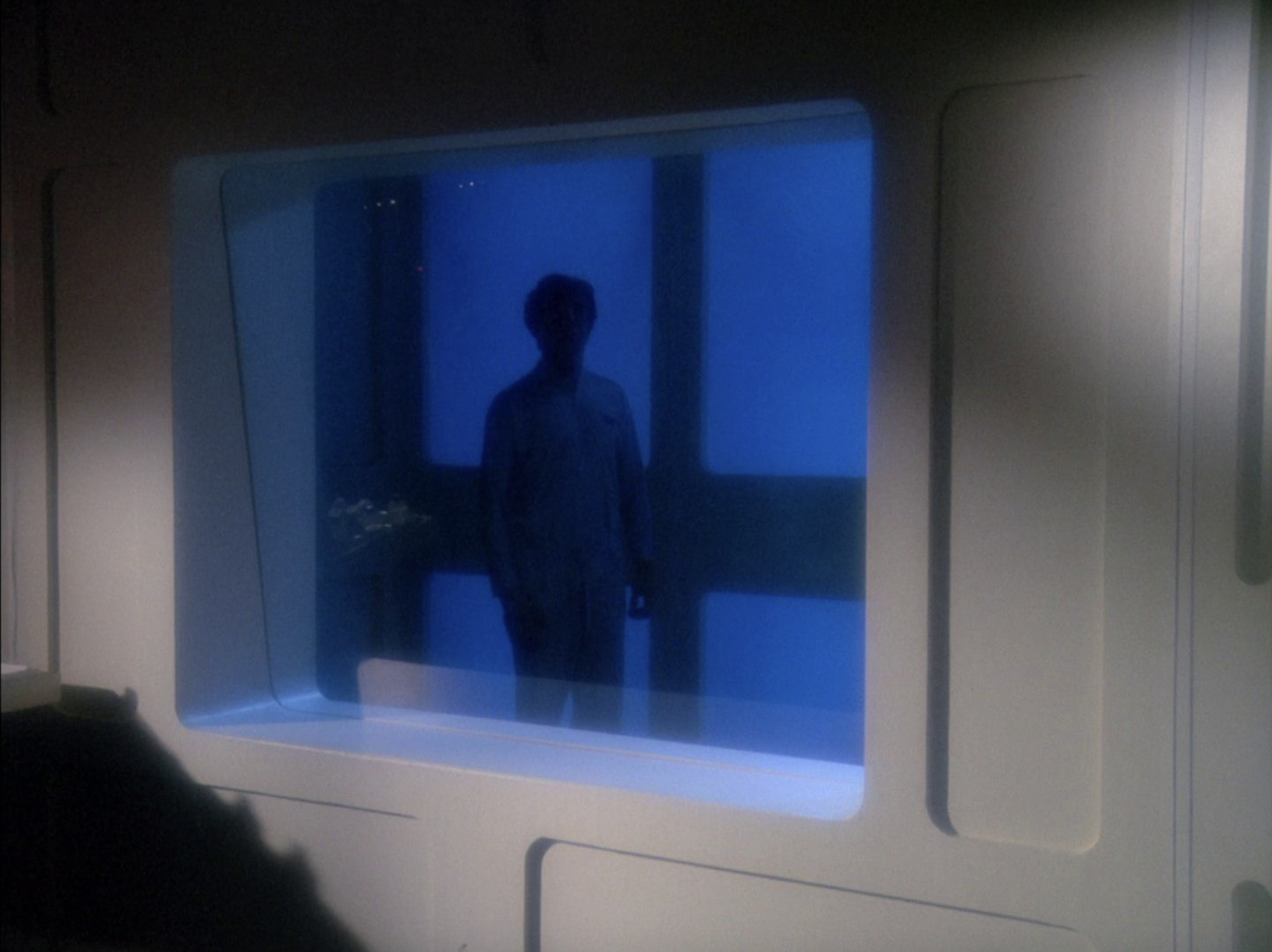 A figure covered by shadow watches through the glass of the sickbay window.