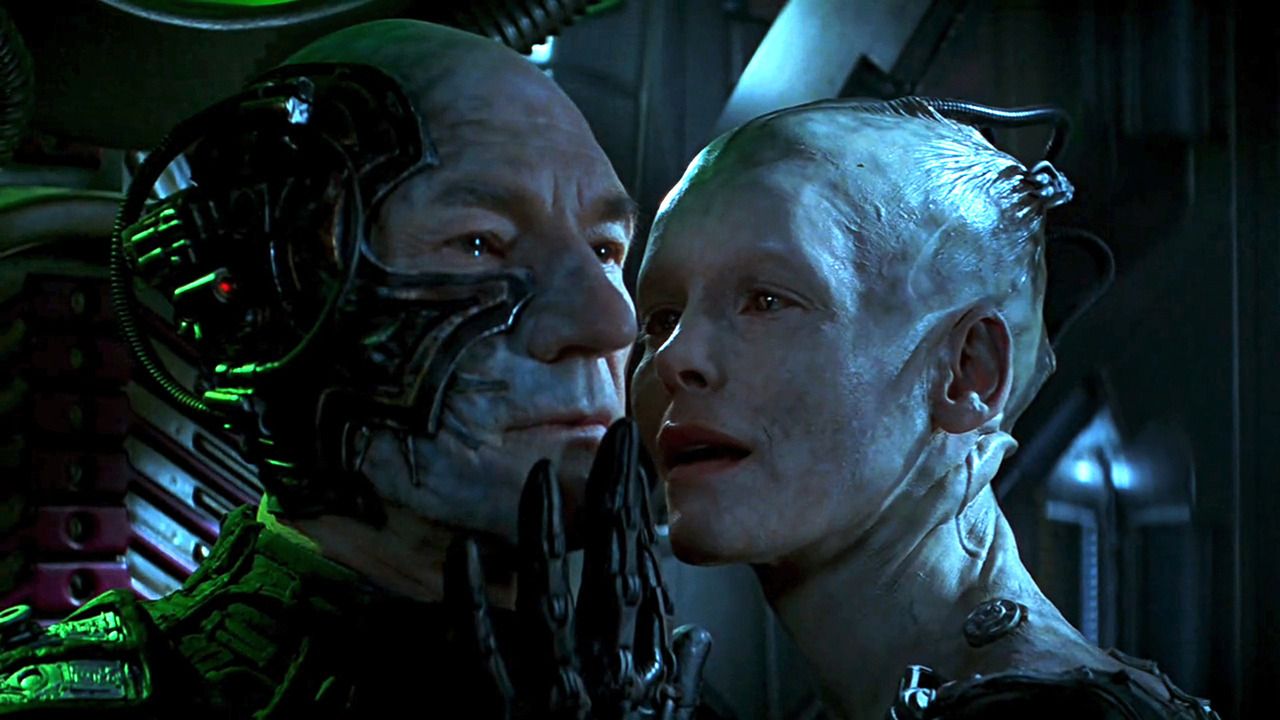 Jean-Luc Picard (Patrick Stewart) as a Borg, half his face obscured by cybernetic implants. The Borg Queen (Alice Krieg) carresses his face.