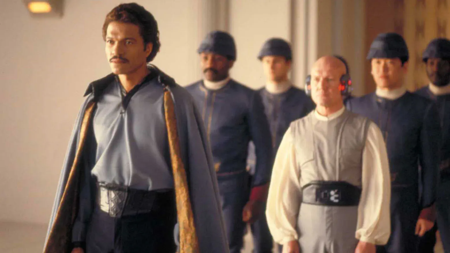 Lando Calrissian (Billy Dee Williams) wears a cloak and is followed by Lobot (John Hollis) and four Cloud City security in blue uniforms.