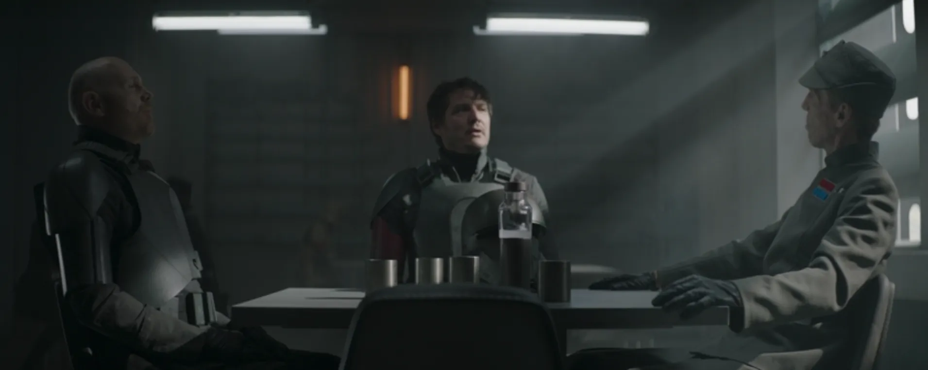 Din Djarin (Pedro Pascal), center, is seated at the center of the table facing the camera with Migs Mayfield (Bill Burr) on his right and an Imperial officer, Valin Hess (Richard Brake) on his left. 