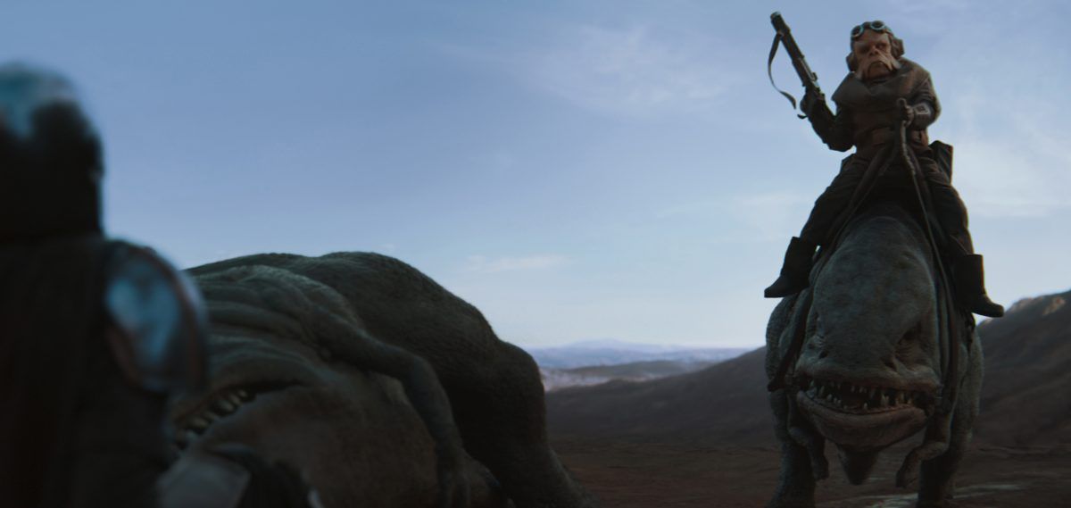 Kuill (voiced by Nick Nolte) holds his blaster up as he greets Mando, visible in the corner of the screen. He is riding a two-legged reptillian creature, with another one slumped to the ground next to him.