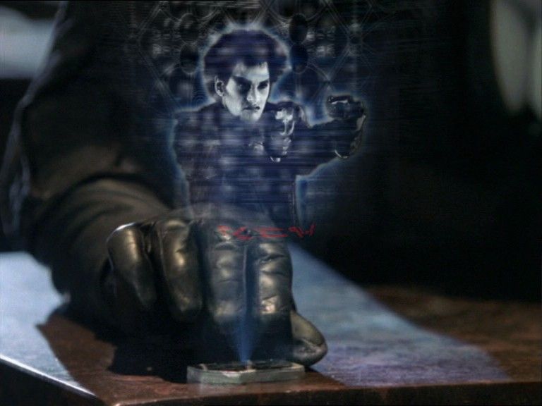 A metal device is placed on a counter, projecting a hologram showing an image of a male Nebari holding two handguns.