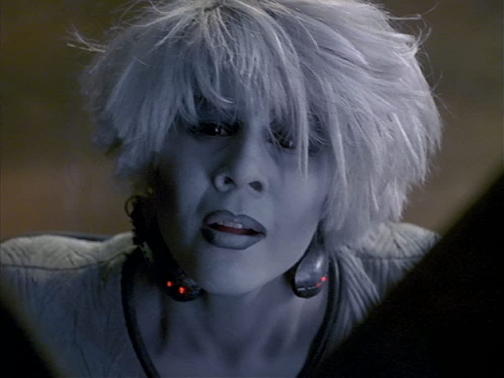 Chiana (Gigi Edgely) wears a glowing collar as she pleads earnestly.