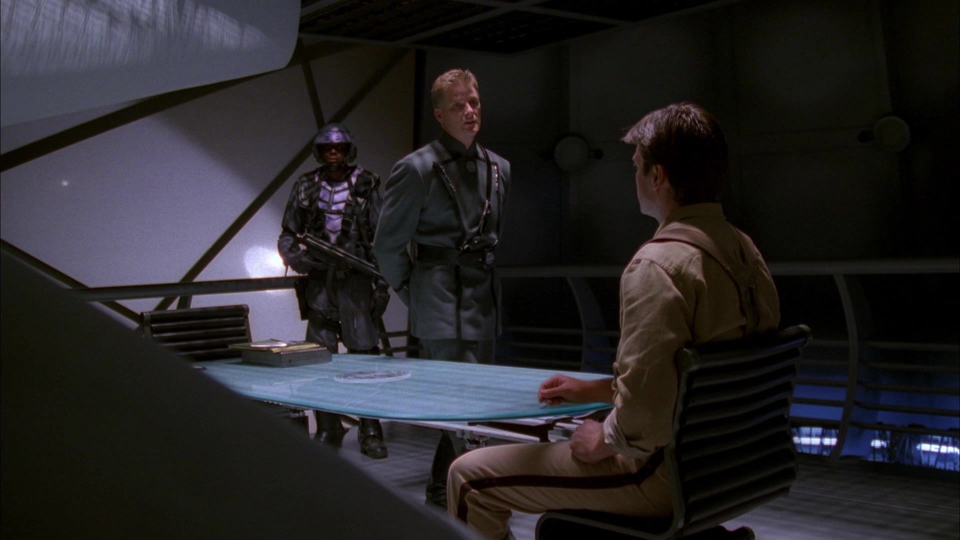 Commander Harken (Doug Savant) stands over Mal Reynolds (Nathan Fillion) in the interrogation room.