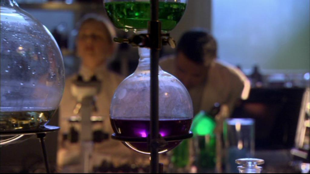 Perna (Allison Hossack) and Dr. Carson Beckett (Paul McGillion) viewed through lab beakers.