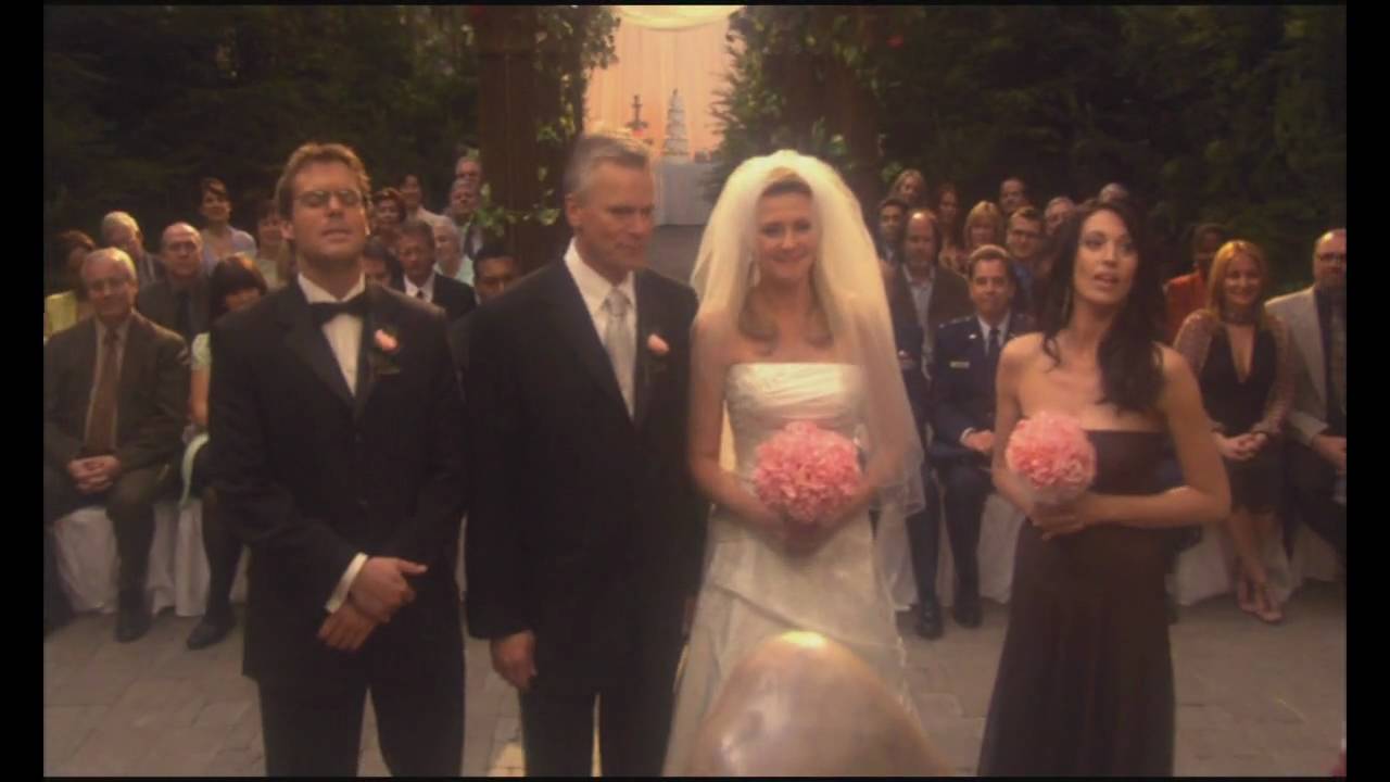 Samantha Carter (Amanda Tapping) in a wedding dress and Jack O’Neill (Richard Dean Anderson) in a suit. Daniel Jackson (Michael Shanks) and Vala Mal Doran (Claudia Black) flank them as best man and maid-of-honor.