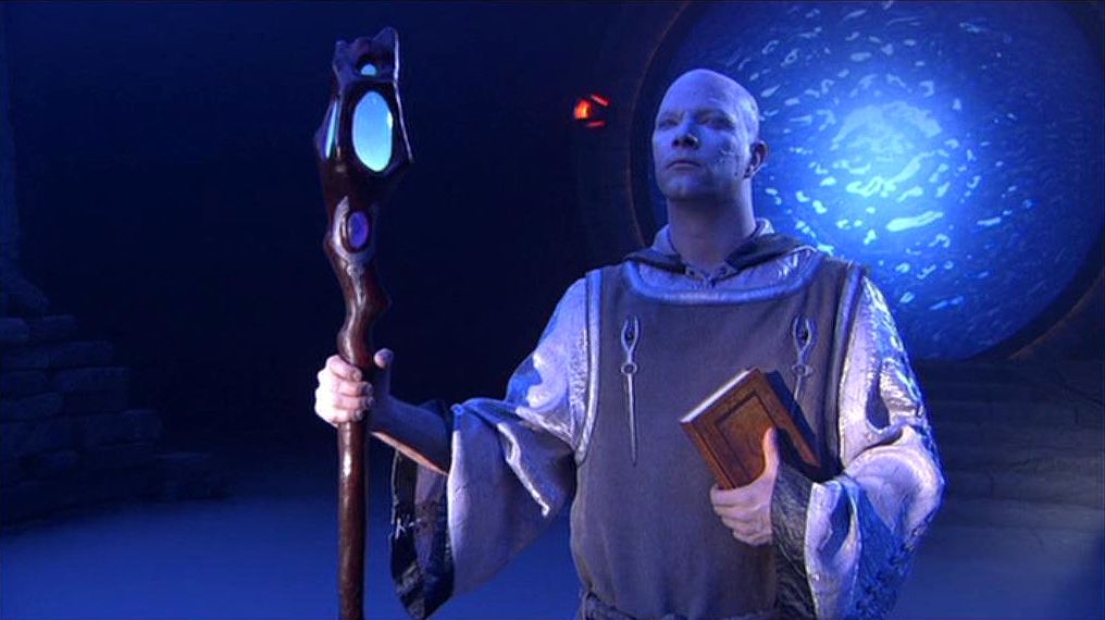 A prior with glowing staff and book in his hand stands in front of the Stargate.