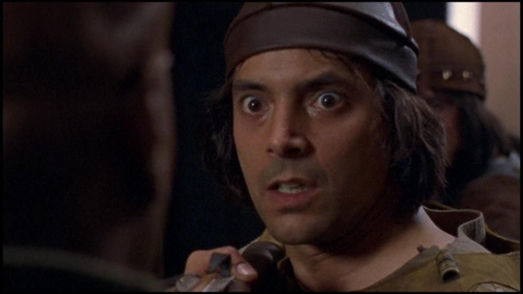 Hanno, a young man with dark shoulder-length hair, addresses Teal'c, who has his back to the camera. Hanno looks angry and has a crossbow-like weapon aimed at Teal'c's chest.