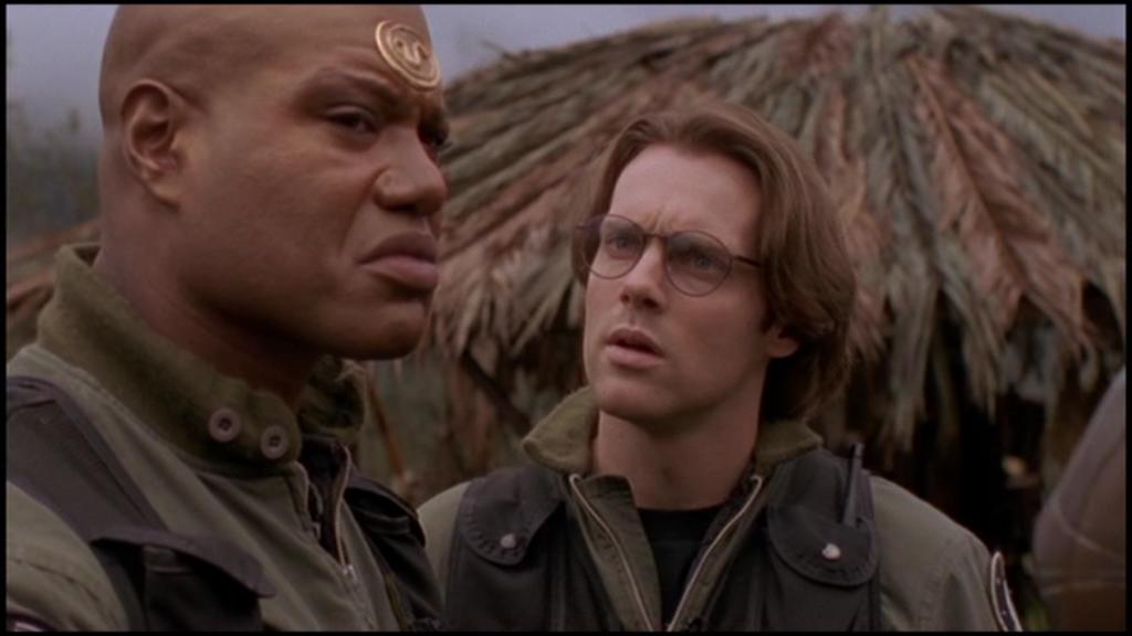 Teal'c looks around warily, Daniel Jackson is asking him a question. In the background the roof of a building made from leaves is visible.