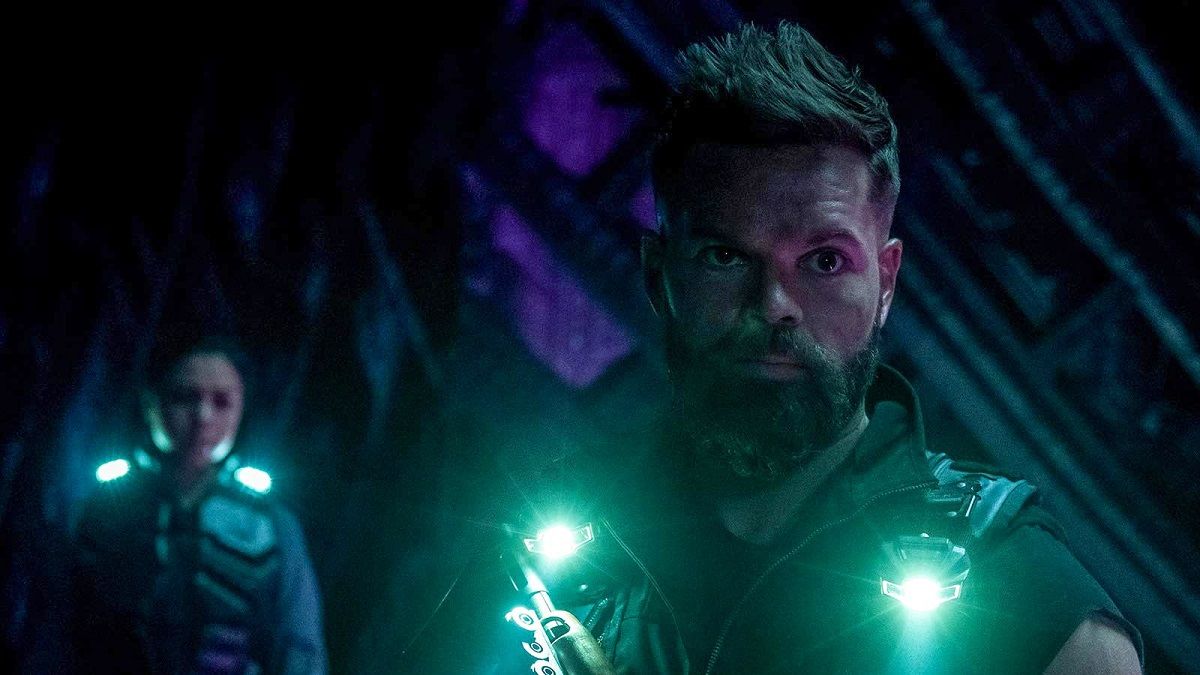 Amos. illuminated by two flashlights pinned to his jacket stands inside the alien structure on Illus. Its walls are grey and patterned. Chandra Wei stands behind him slightly out of focus.