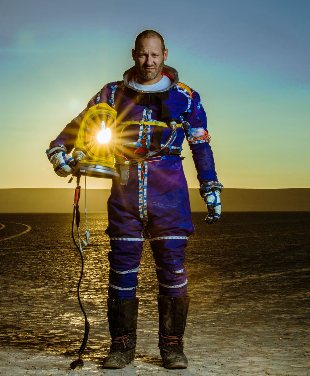 Cameron M. Smith stands in the desert with the light of the sun visible through the helmet under his arm.