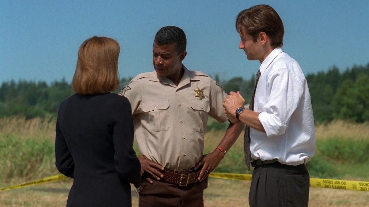 Sheriff Andy Taylor, played by Tucker Smallwood, stands in a field with police tape in the background in The X-Files episode ‘Home’ (S4, Ep2). He is in conversation with Mulder and Scully.