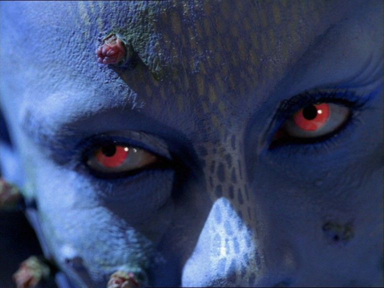 A close-up of Zhaan’s face, her eyes red, and her skin marked with buds.
