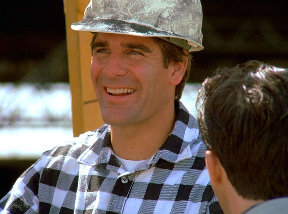 Sam (Scott Bakula) grins, he wears a checked shirt and hard hat.