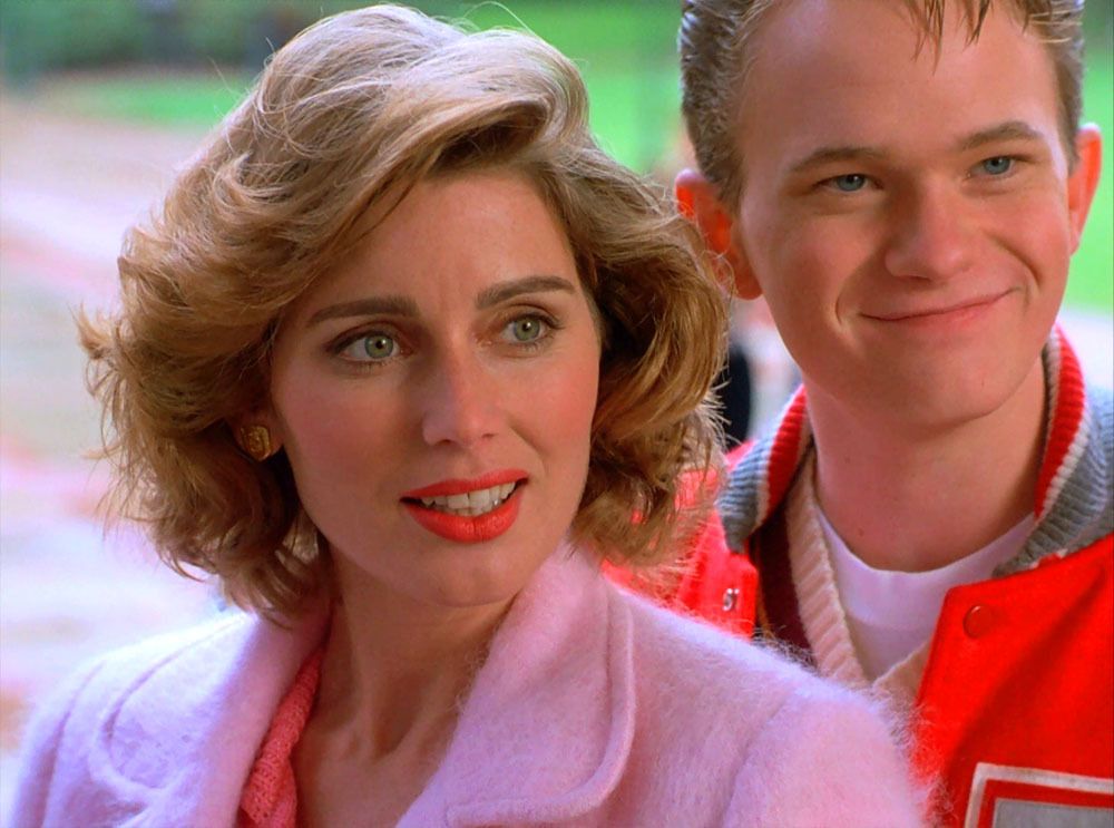 Alia (Renée Coleman) wears a pink jacket and bright red lipstick, whilst Mike (Neil Patrick Harris) stands beside her in a red letterman jacket.