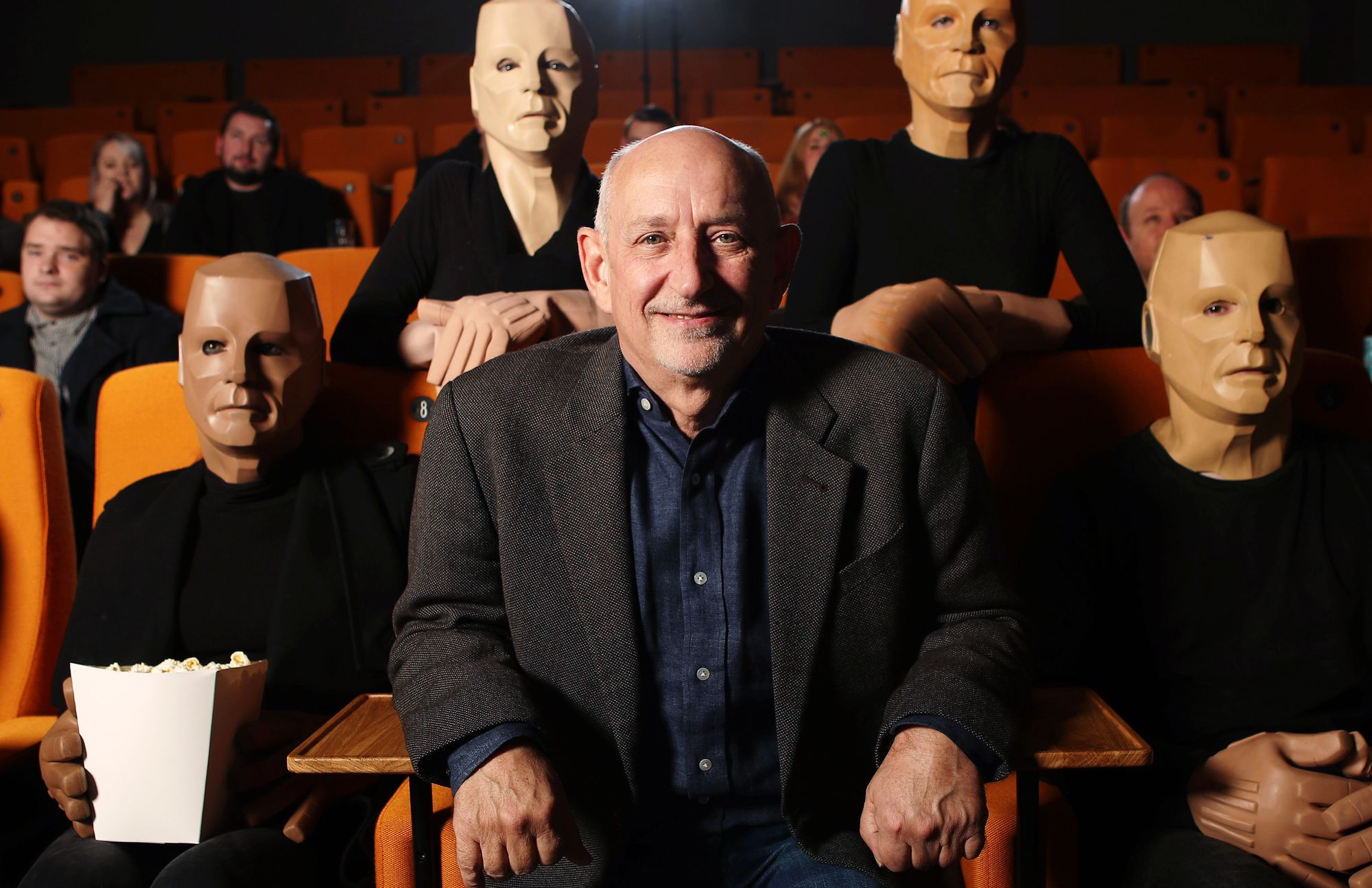 Doug Naylor sits in cinema stalls surrounded by figures in Kryten-style masks.