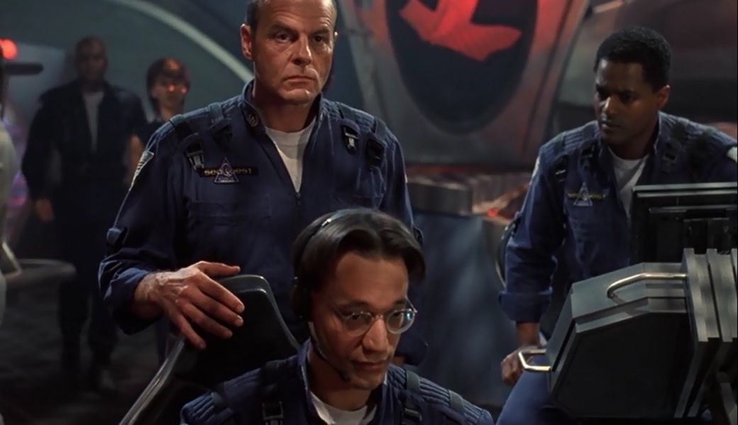 Captain Oliver Hudson (Michael Ironside) stands behind Lt. Timothy O’Neil (Ted Raimi), with his hand on the back of his chair.
