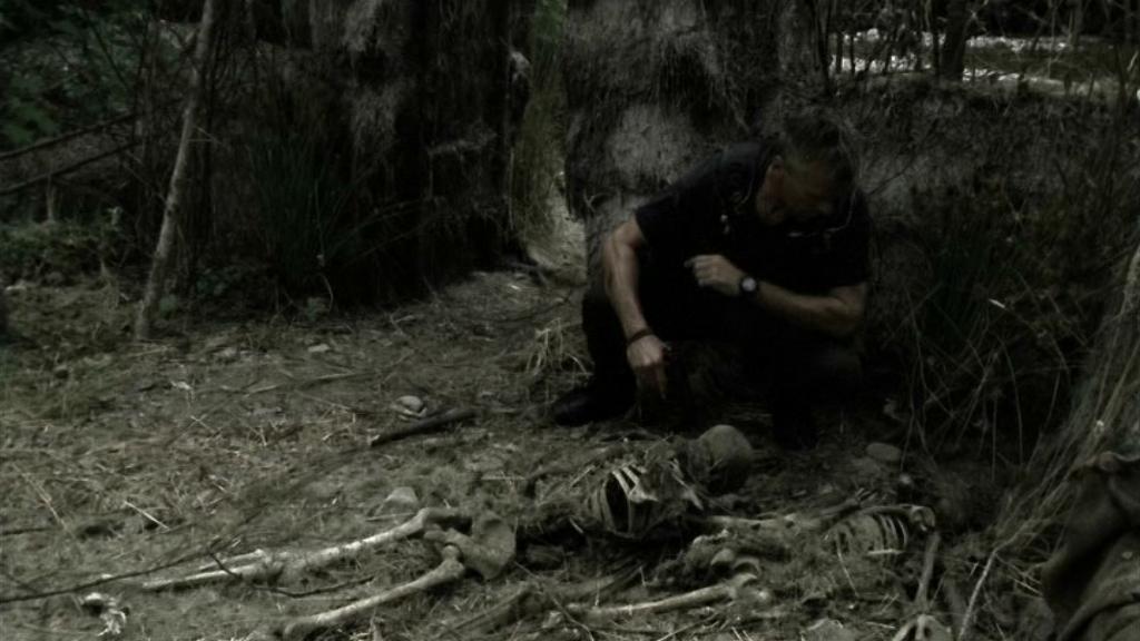 Jack O’Neill (Richard Dean Anderson) crouches next to two human skeletons.