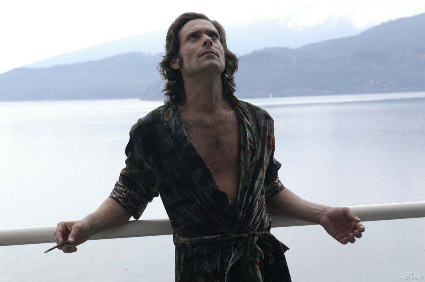 Gaius Baltar (James Callis) leans against a rail with his palms up and his face turned upwards.