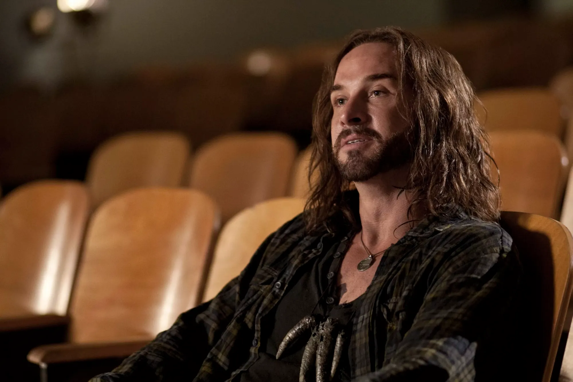 John Pope (Colin Cunningham) in the Falling Skies episode ‘The Armory’. Pope, who has long hair, and wears an open plaid shirt with a medallion and a necklace of alien fingers beneath is sitting in theater stalls addressing someone off-camera.