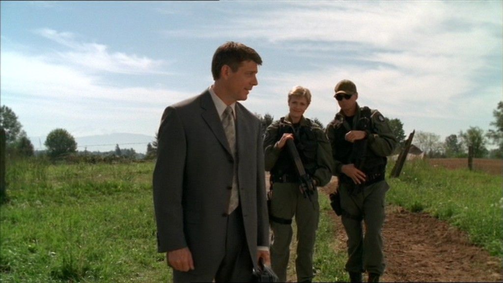 Ambassador Joe Faxon (Christopher Cousins), Samatha Carter (Amanda Tapping), and Jack O’Neill (Richard Dean Anderson) walk across farmland.