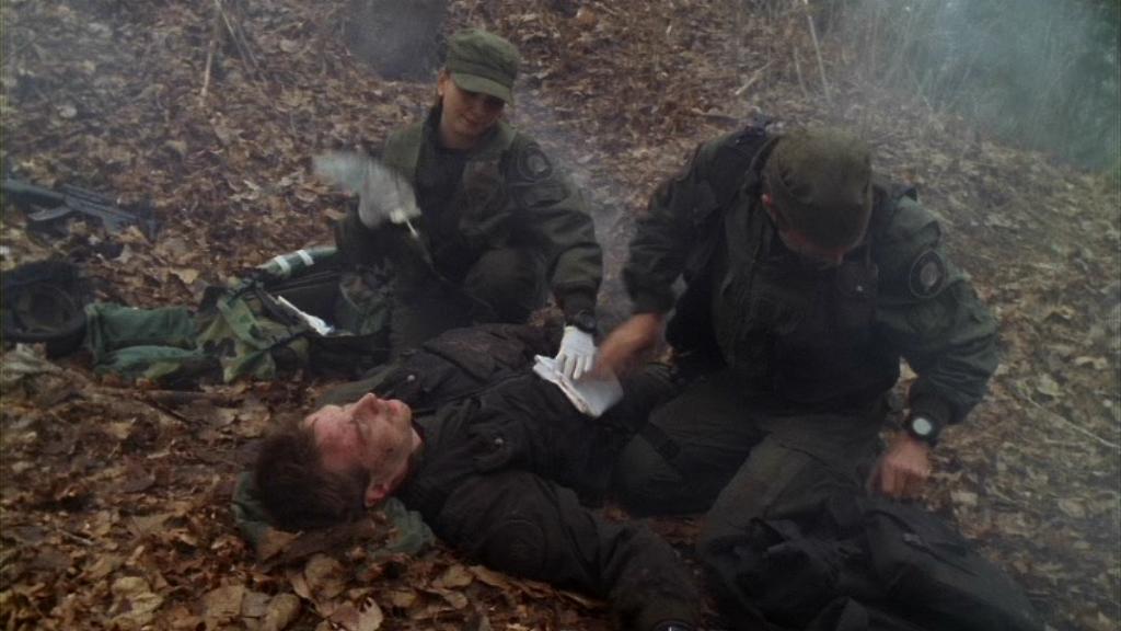 Janet Fraiser (Teryl Rothery) and Daniel Jackson (Michael Shanks) tend to the wounded Senior Airman Simon Wells (Julius Chappel) in the Stargate SG-1 episode ‘Heroes, Part 2’. Daniel is holding dressing over a wound in the soldier's torso, whilst Janet has her hand on his shoulder. They are on the floor in a forest, the ground covered in dead leaves.