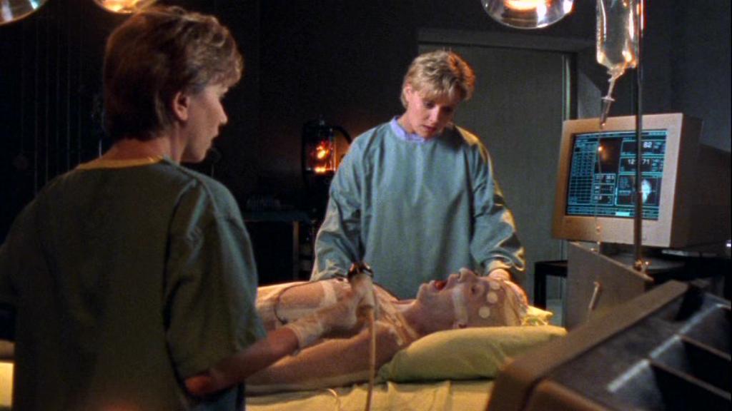 Samantha Carter, played by Amanda Tapping, examines one of the aliens in the Stargate SG-1 episode 'One False Step'. Samantha is wearing blue surgical scrubs and the alien is lying on a hospital bed in a lab in Stargate Command.