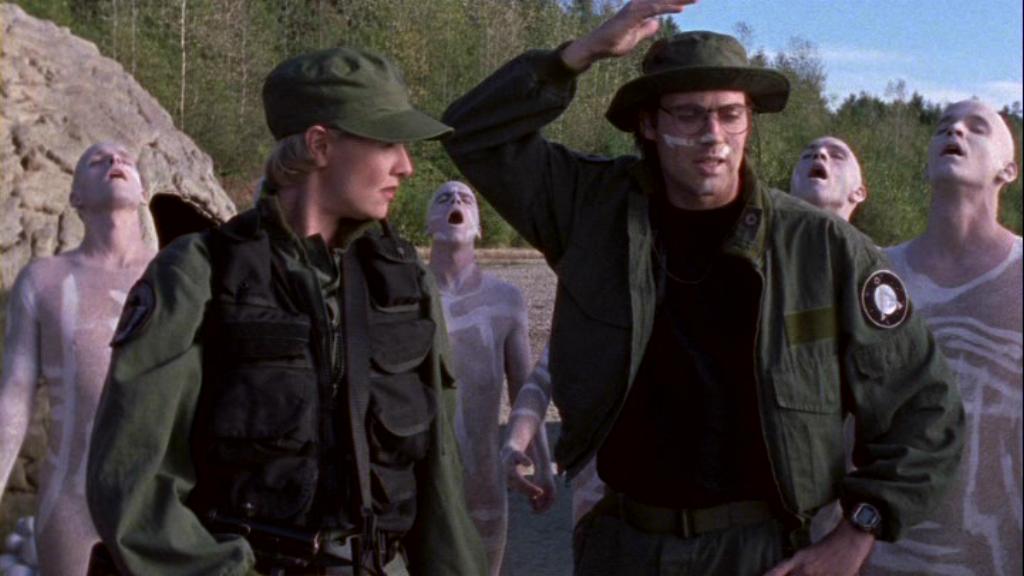 Samantha Carter, played by Amanda Tapping, and Daniel Jackson, played by Michael Shanks, in Stargate SG-1 episode 'One False Step'  (S2, Ep19). | MGM, 1999. Samantha and Daniel are surrounded by bald men who are singing with their heads tipped back and their eyes closed. They are seemingly naked and marked with white paint.