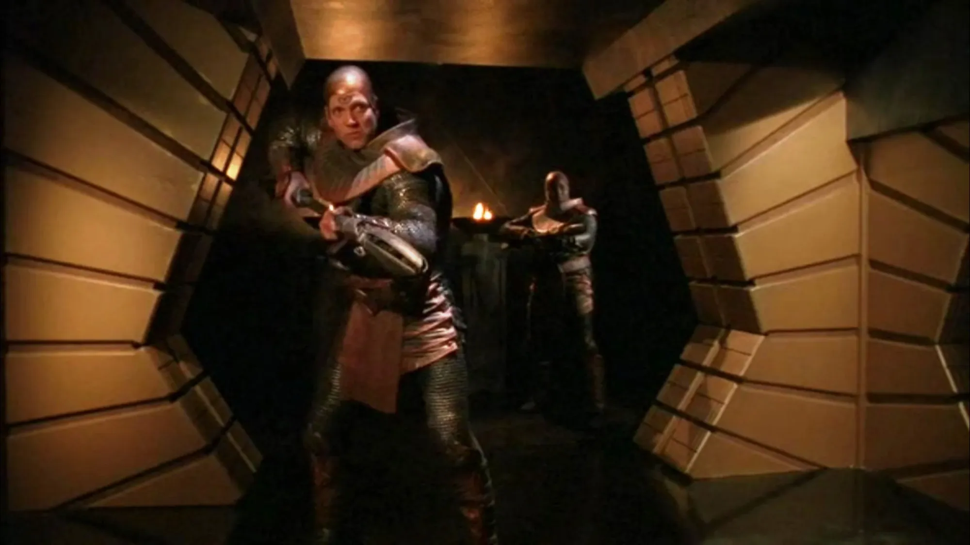 Two Jaffa warriors in golden armor with staff weapons in the Stargate SG-1 episode ‘Upgrades'. They are standing in the corridor of a ship with their staffs ready to fire.