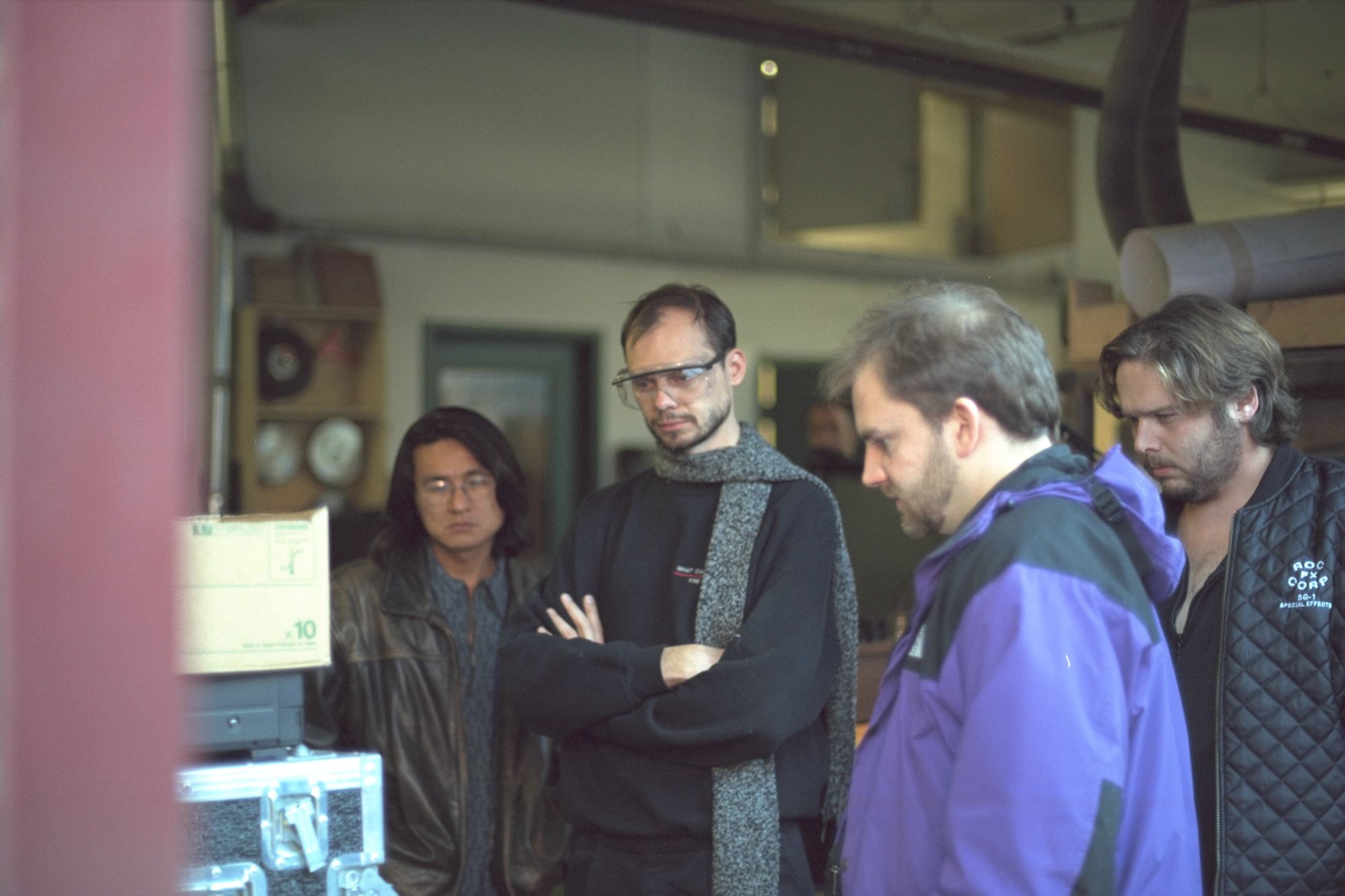 A group of men stand around a monitor in a VFX workshop of some kind. John Gajdecki is wearing safety goggles.ss