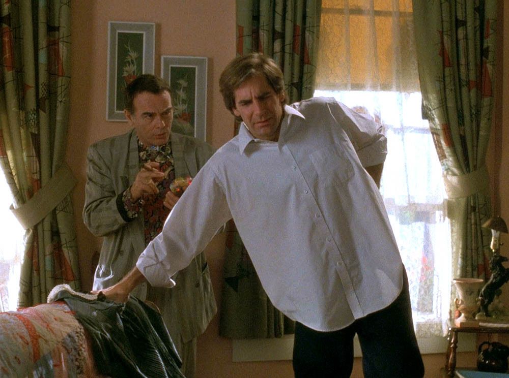 A pregnant Sam Beckett (Scott Bakula) leans on the sofa and rubs his lower back.