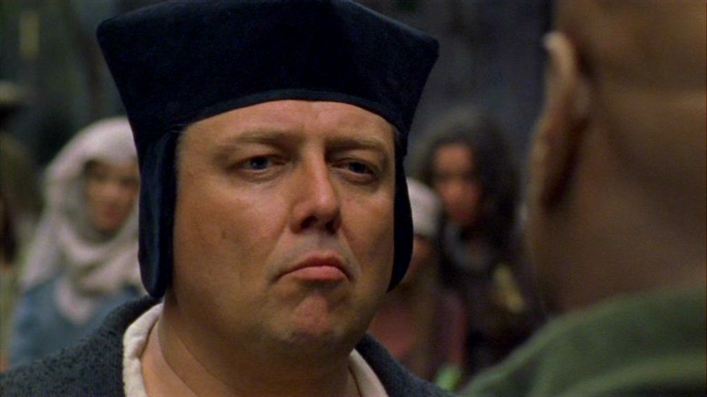 The Canon, played by A.C. Peterson, pulling a sneer in  in the Stargate SG-1 episode 'Demons'. Behind him, a soldier stands holding a spear.